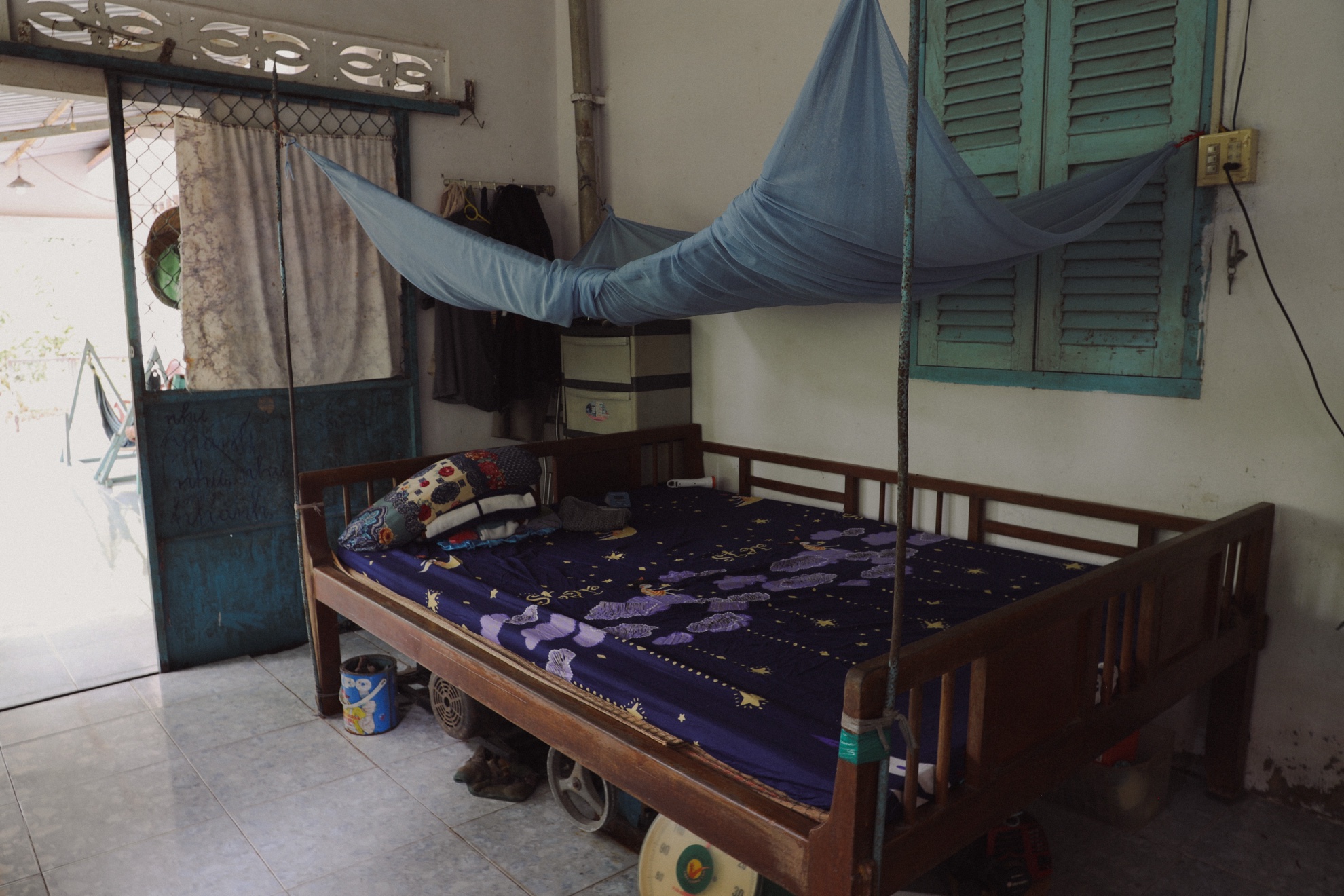 A wooden bed under a blue mosquito net, the front door open to the yard.