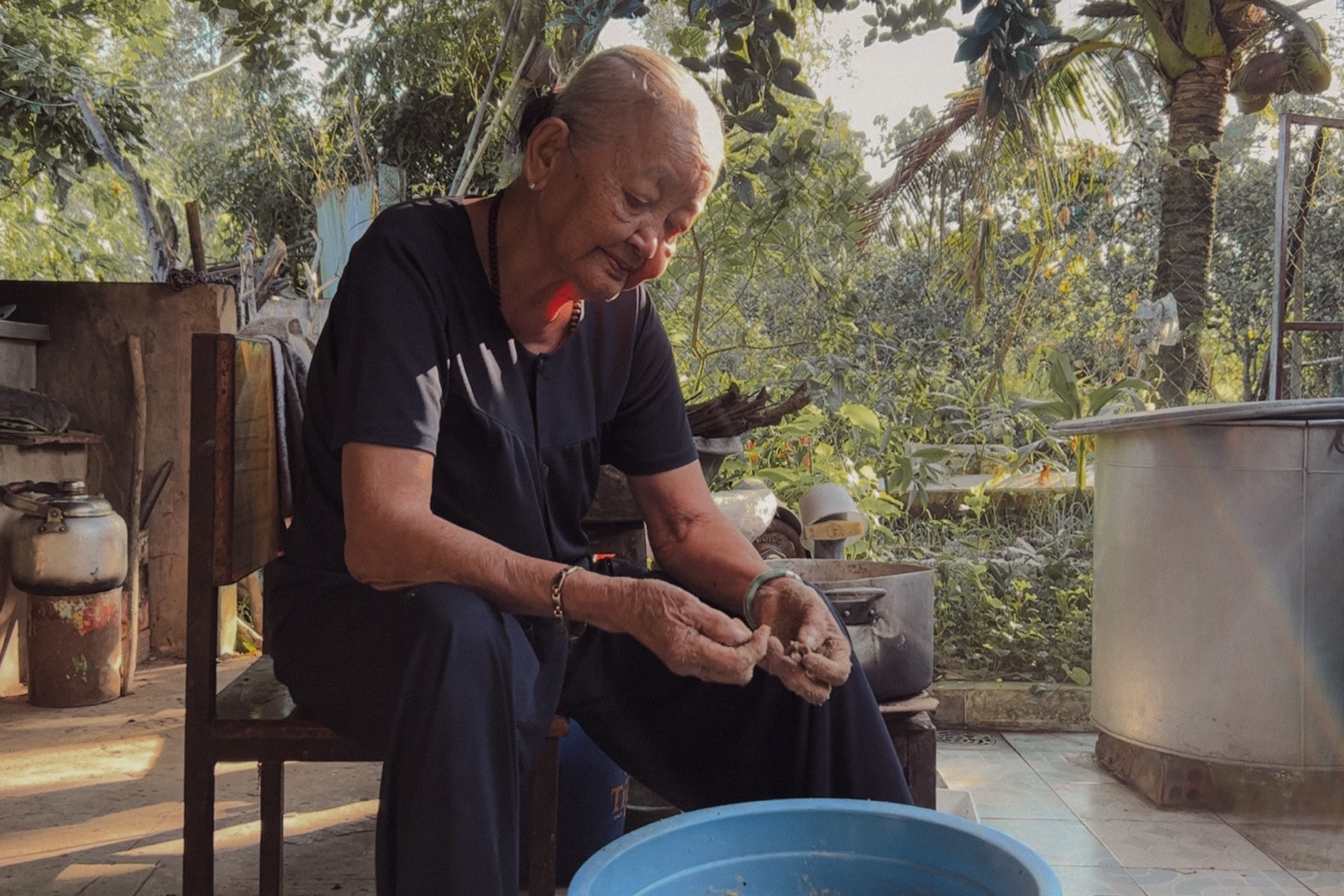 Grandmother seated in the yard, working over a blue basin with her hands.
