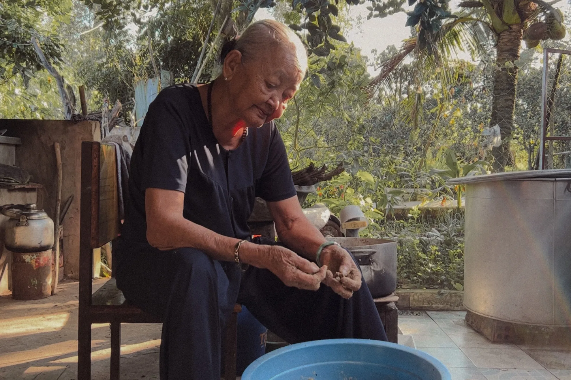 Grandmother seated in the yard, working over a blue basin with her hands.