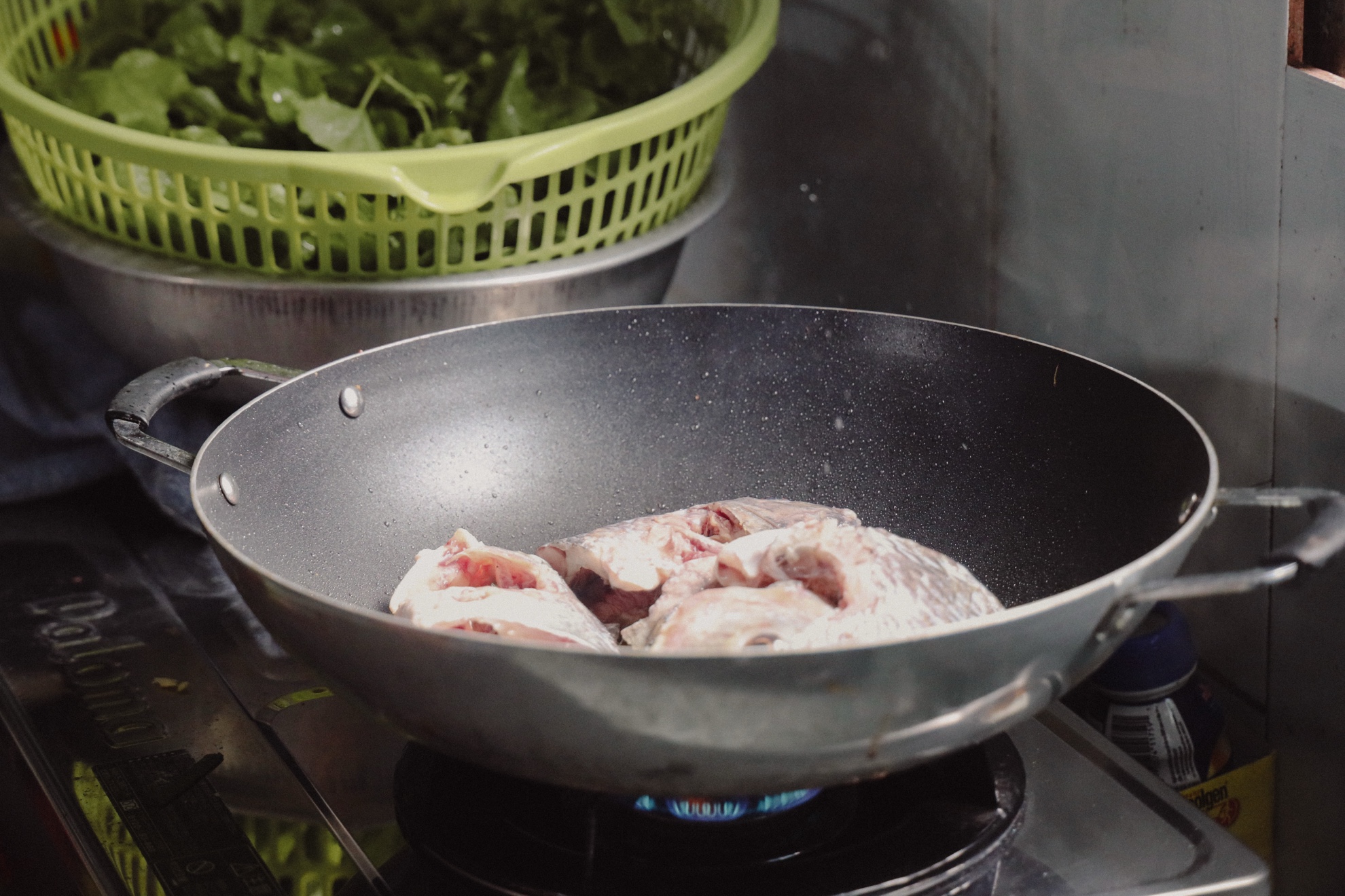 Fish in a wok over a blue gas flame, a basket of greens behind.